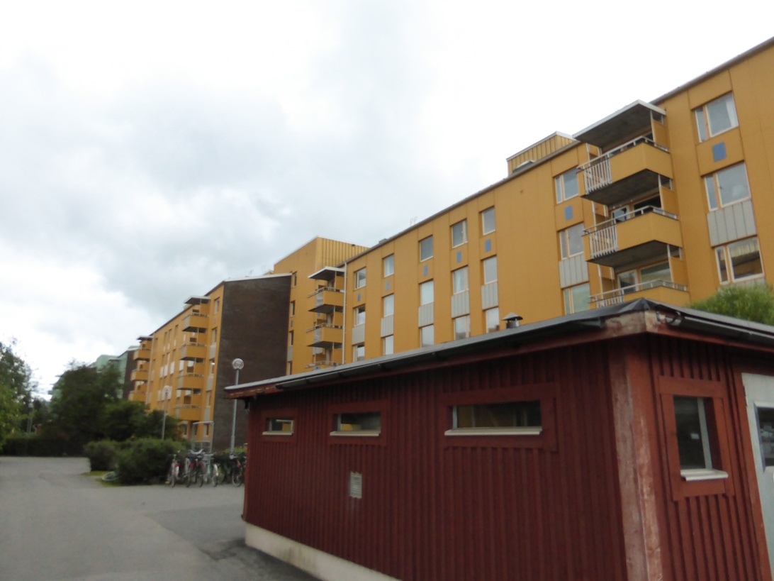 Ett gult flervåningshus med balkonger i ett bostadsområde i Stockholm, med en liten röd komplementbyggnad i förgrunden.