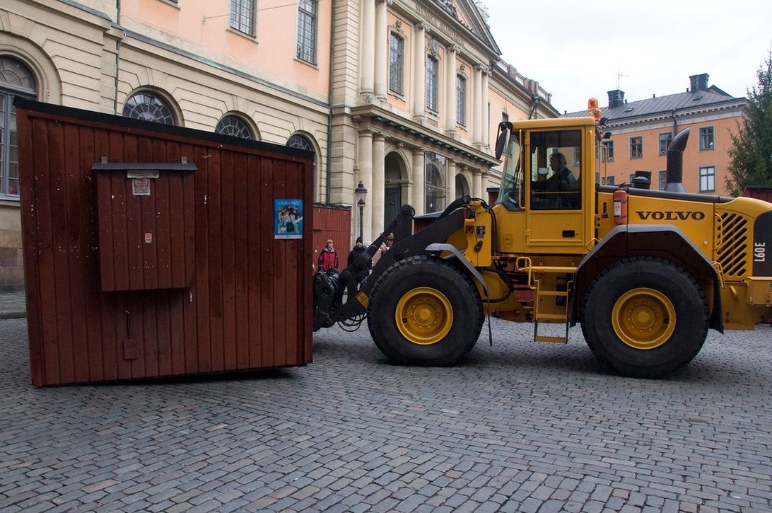 En gul Volvo hjullastare transporterar en träbod på en kullerstensgata i Stockholm, med historiska byggnader i bakgrunden.
